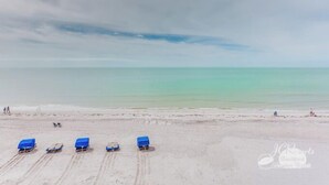 Una playa con varias sillas azules dispuestas en la arena, algunas personas a lo lejos y un cielo nublado.