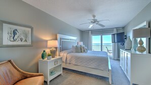 A bedroom with a bed, nightstands, a chair, a ceiling fan, a TV, and a large window.