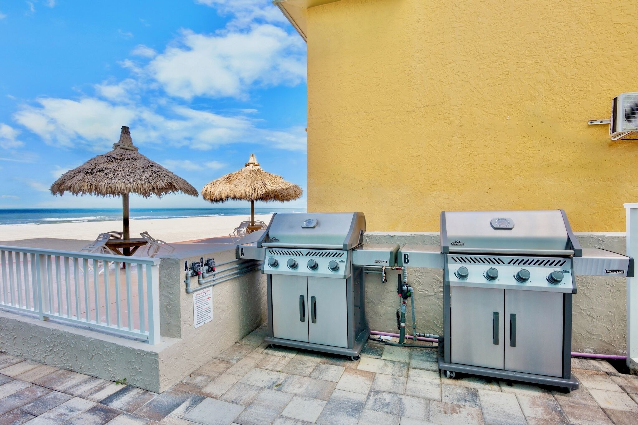 Een terras met twee barbecue grills, een rieten zonnescherm en uitzicht op het strand.