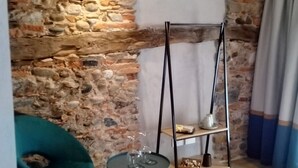 Habitación de esquina con pared de piedra, una silla color turquesa, una pequeña mesa redonda con vasos y una estantería metálica.