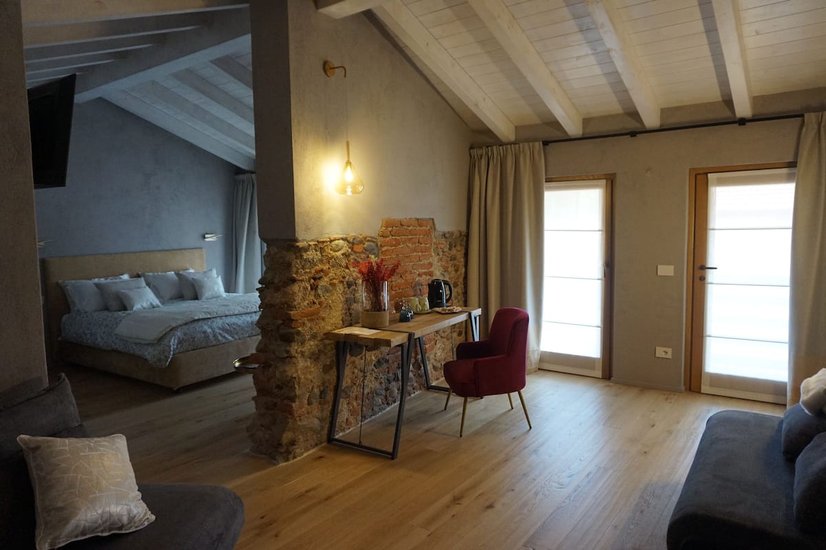A cozy room with a bed, a red chair, a small table with a vase, and a stone wall.