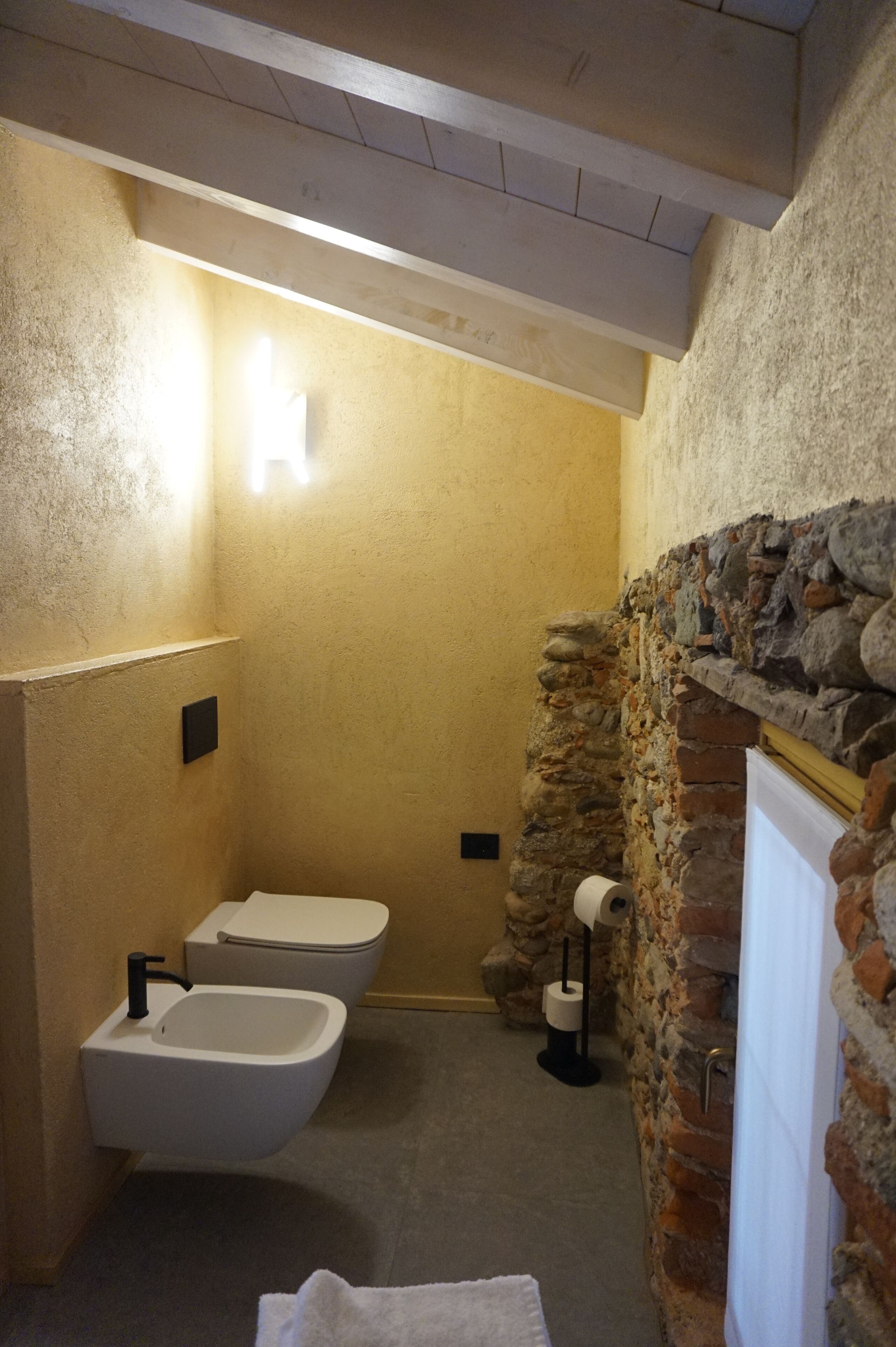 Un baño con pared de piedra, un lavamanos blanco, dos inodoros y una toalla en el suelo.