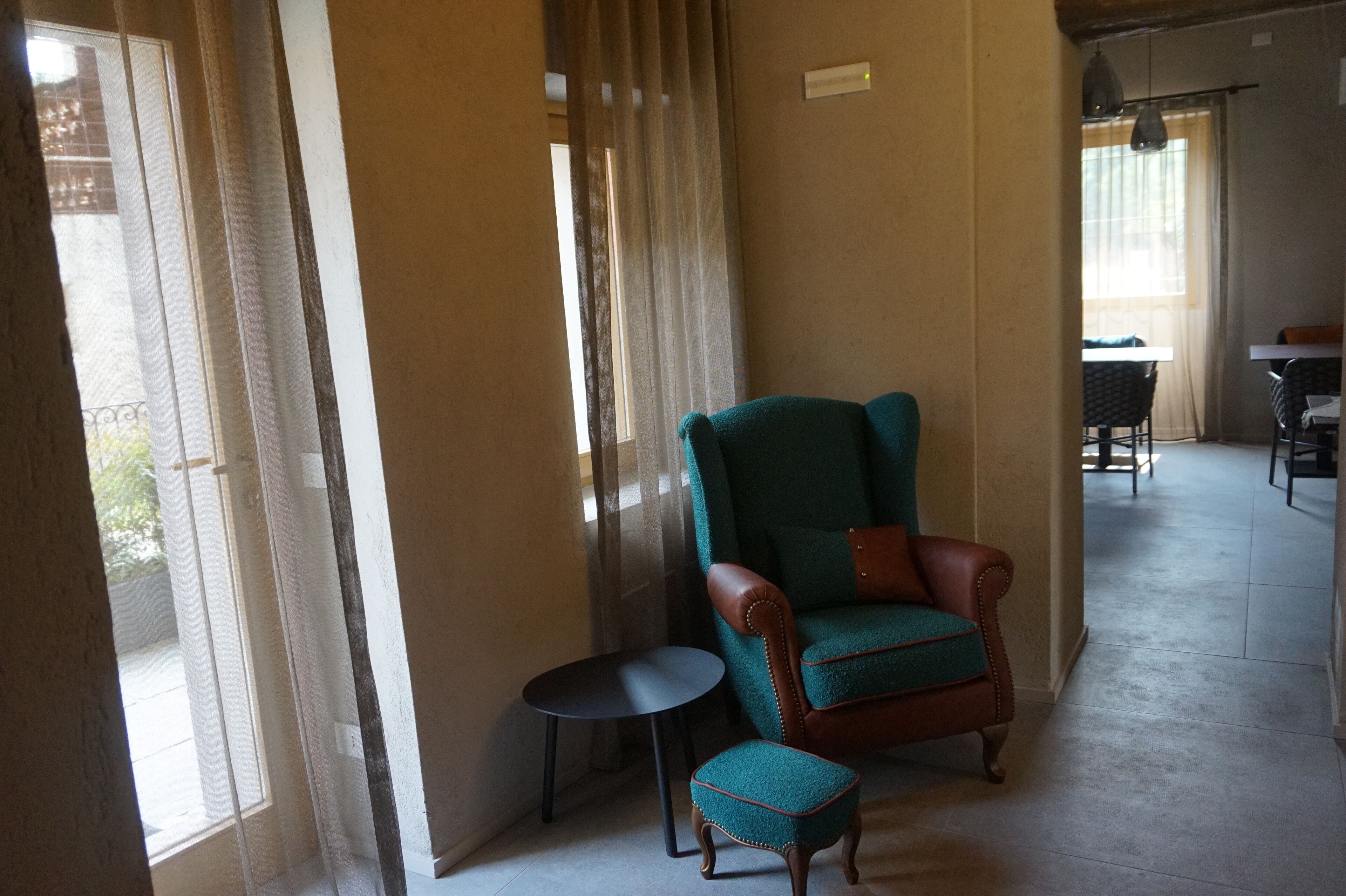 Quarto com uma janela grande, uma poltrona verde-água, uma mesa redonda pequena e um banquinho combinando.