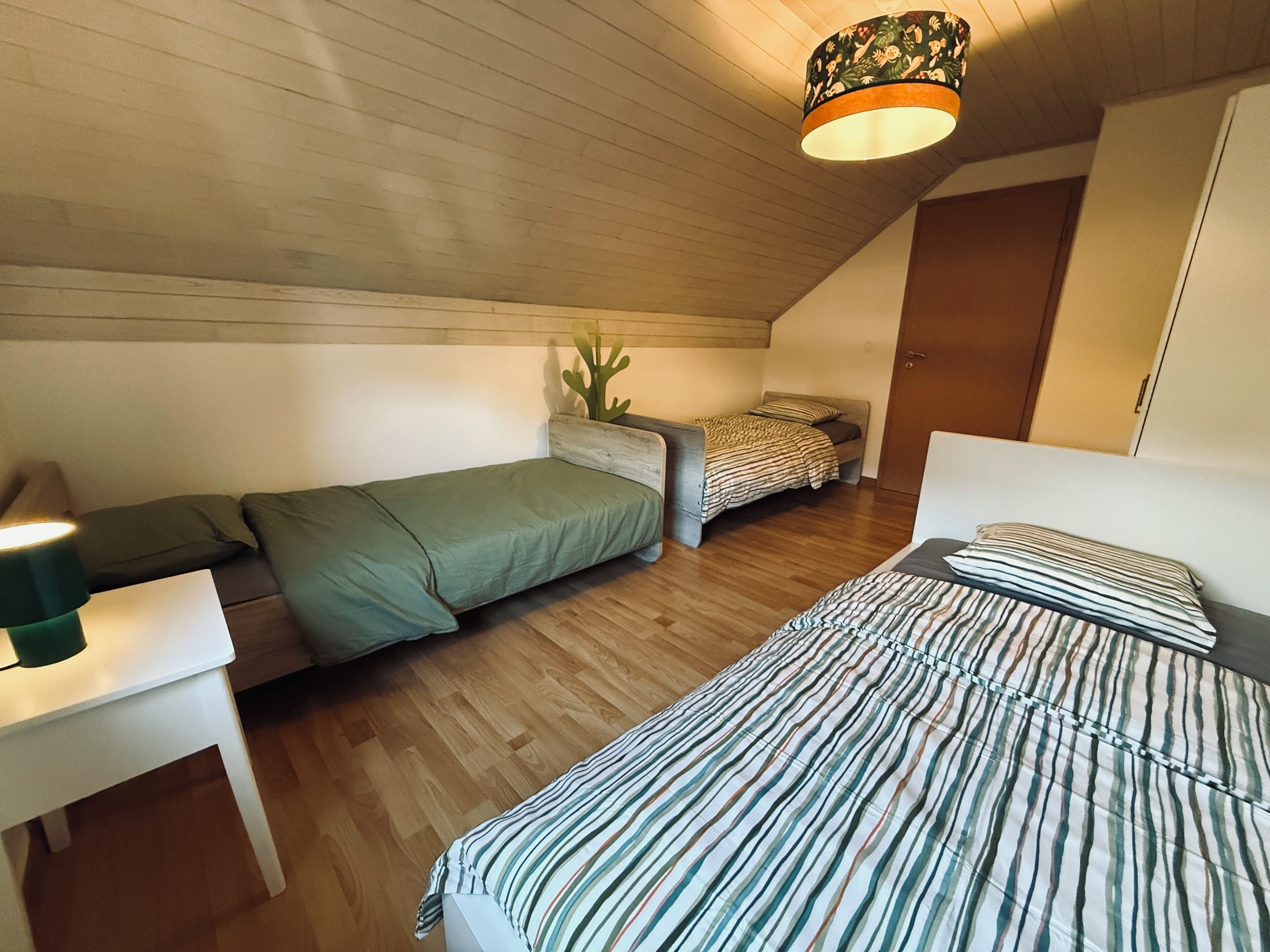 Ein kleiner, gemütlicher Raum mit schrägem Dach, Holzboden, einem Bett mit gestreifter Bettwäsche, einem kleinen Tisch mit einer Lampe und einer Kakteenpflanze.
