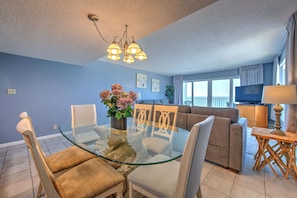 A modern living room with a glass dining table, beige chairs, a sofa, and a television.