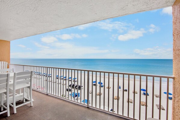 A balcony with a view of a beach, white chairs, and blue umbrellas.