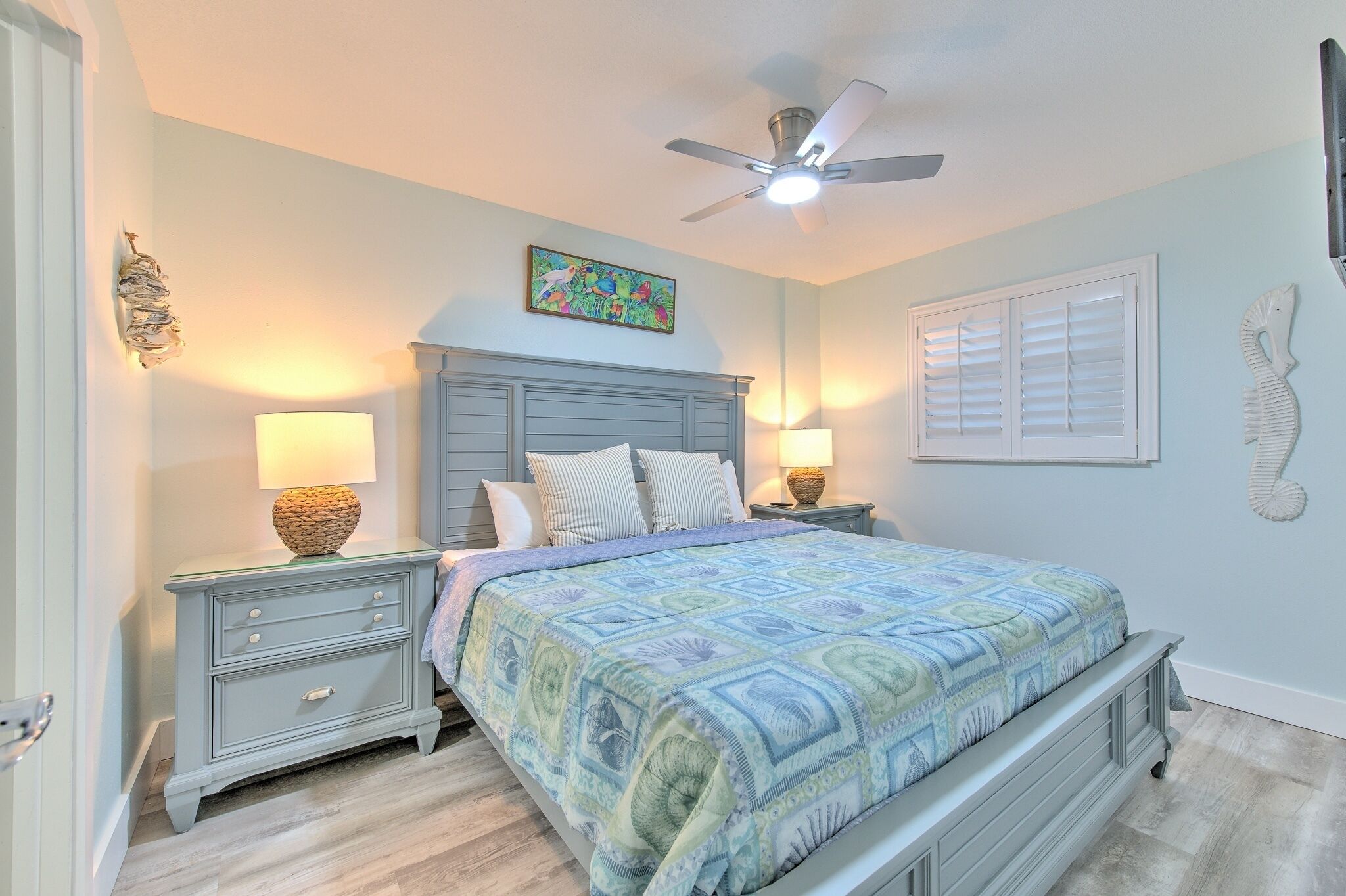 A bedroom with a bed, two bedside tables with lamps, a ceiling fan, a window with shutters, and a decorative seahorse on the wall.