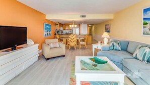 A living room with a TV, sofa, and coffee table. There is a dining area with chairs and a table. The room has orange walls and a ceiling light.
