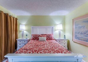 A bed with a patterned bedspread, two bedside tables with lamps, a nightstand, a framed picture on the wall, and a window with curtains. - Top Floor Gulf Front Huge Condo w/ Balcony RS506 (N. Redington Beach)