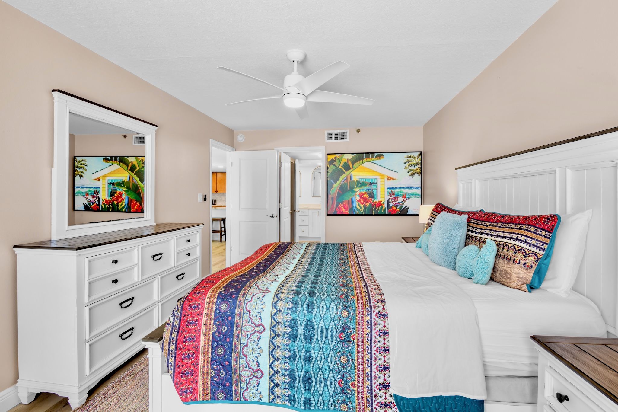 A bedroom with a bed, a dresser, a ceiling fan, and two paintings on the wall.