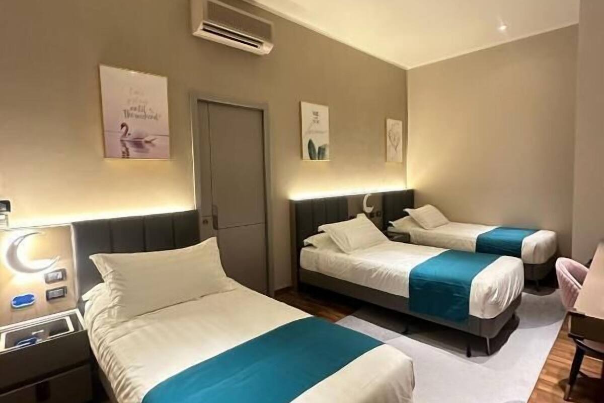 A hotel room with two beds, a desk, a chair, and a small table. There is an air conditioning unit on the wall and a door.