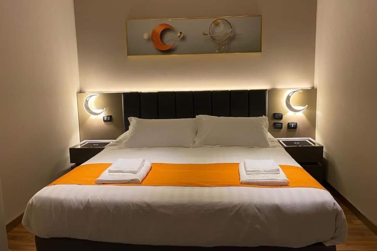 A neatly made bed with white linens and an orange accent, a modern headboard, and wall-mounted nightstands with ambient lighting.
