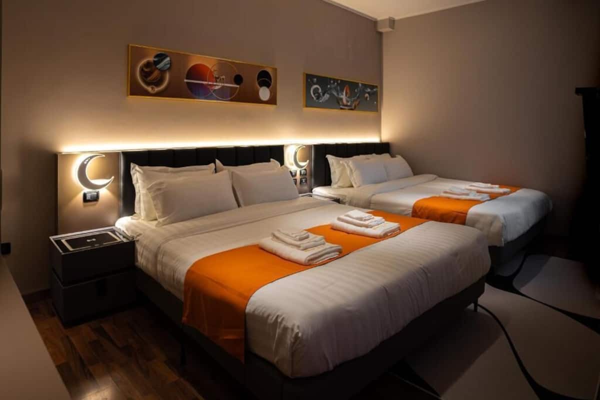 A hotel room with two beds, each with white bedding and orange accents, a nightstand with a lamp, and artwork on the walls.