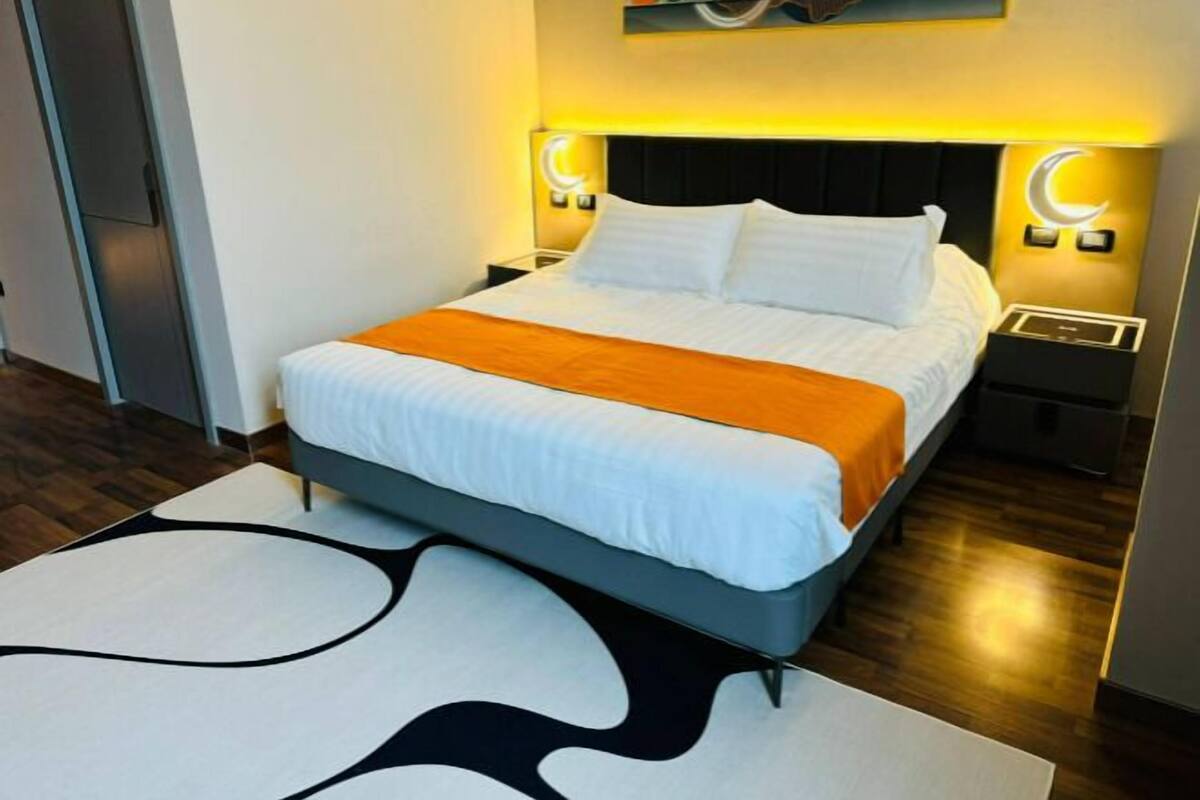 A modern hotel room with a neatly made bed, a large abstract painting on the wall, and a unique black and white rug.