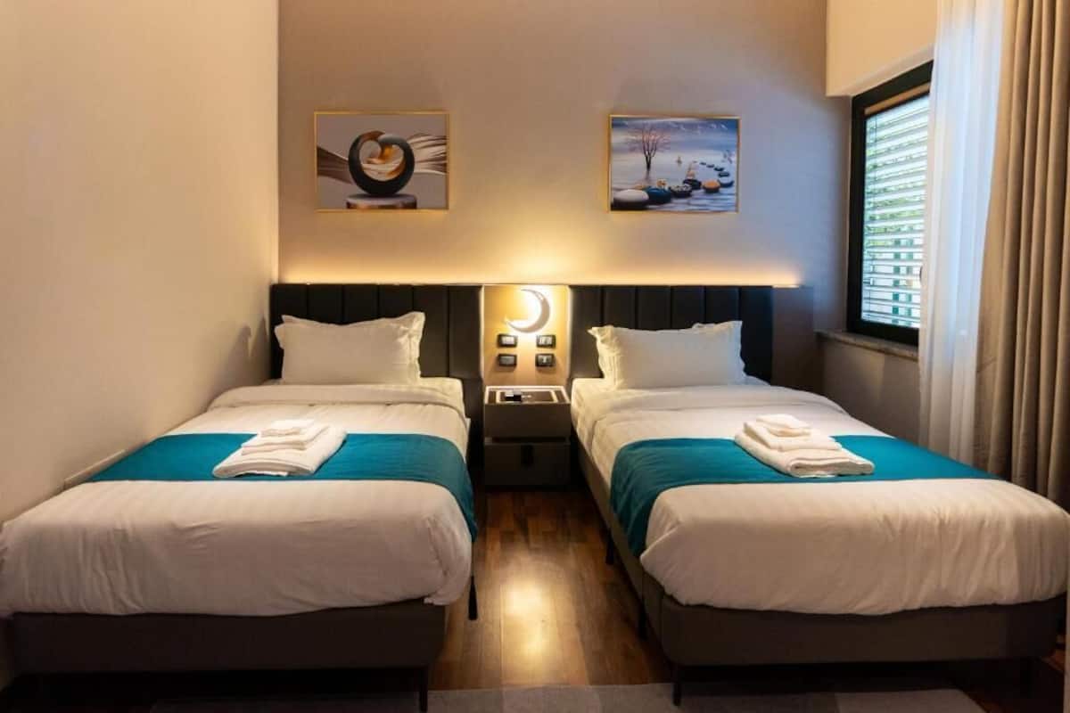 A hotel room with two beds, each with white bedding and teal accents, a nightstand with a crescent moon light, and artwork on the walls.