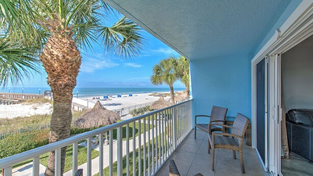 A balcony with a view of the beach, palm trees, and a thatched structure.