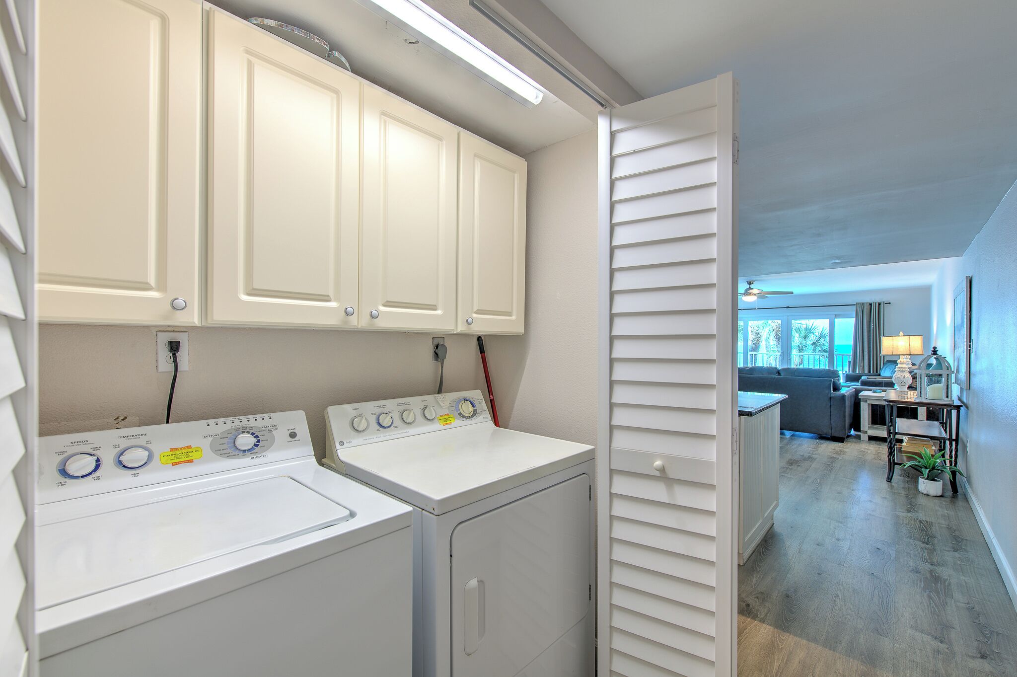 A laundry area with two washing machines, a cabinet above, and a view into a living room with a sofa and a table.