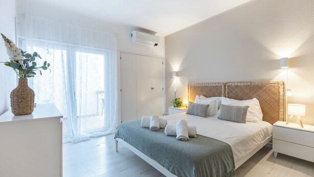 A bedroom with a bed, bedside tables, a nightstand, a window with curtains, a wall-mounted air conditioner, and a vase with flowers.