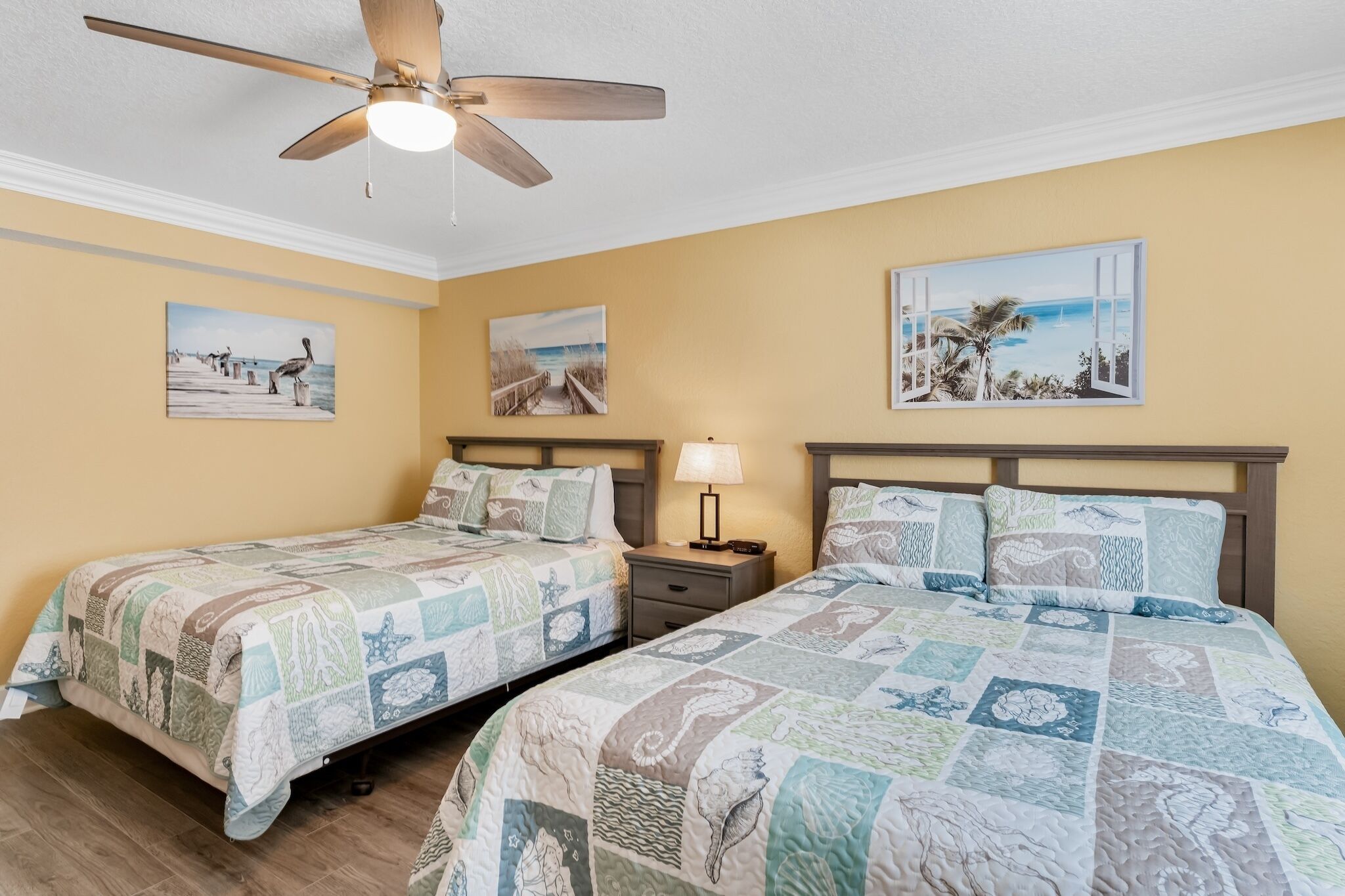 A bedroom with two beds, a ceiling fan, and wall art depicting coastal scenes.