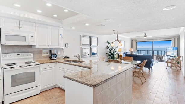 A modern kitchen with white appliances, a granite countertop, and a dining area with a view of the ocean. - Light and Airy Gulf Front Condo w/ Balcony - SD205 (Indian Shores)