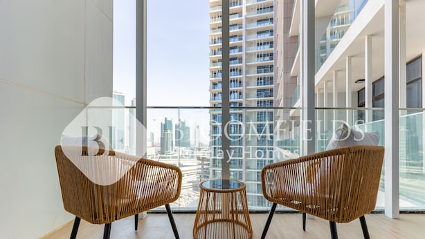 Un soggiorno moderno con due poltrone in vimini e un tavolino rotondo, ampie finestre con vista sulla città e un balcone con ringhiere in vetro.