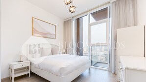 A modern bedroom with a large bed, a nightstand, a bedside lamp, a window with curtains, and a city view. - Bloomfields Spacious 1BR Bloom Tower (Dubai)