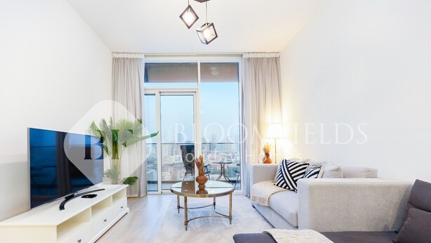 A modern living room with a sofa, a coffee table, and a TV on a stand. There is a large window with curtains, a plant, and a city view. - Bloomfields Spacious 1BR Bloom Tower (Dubai)