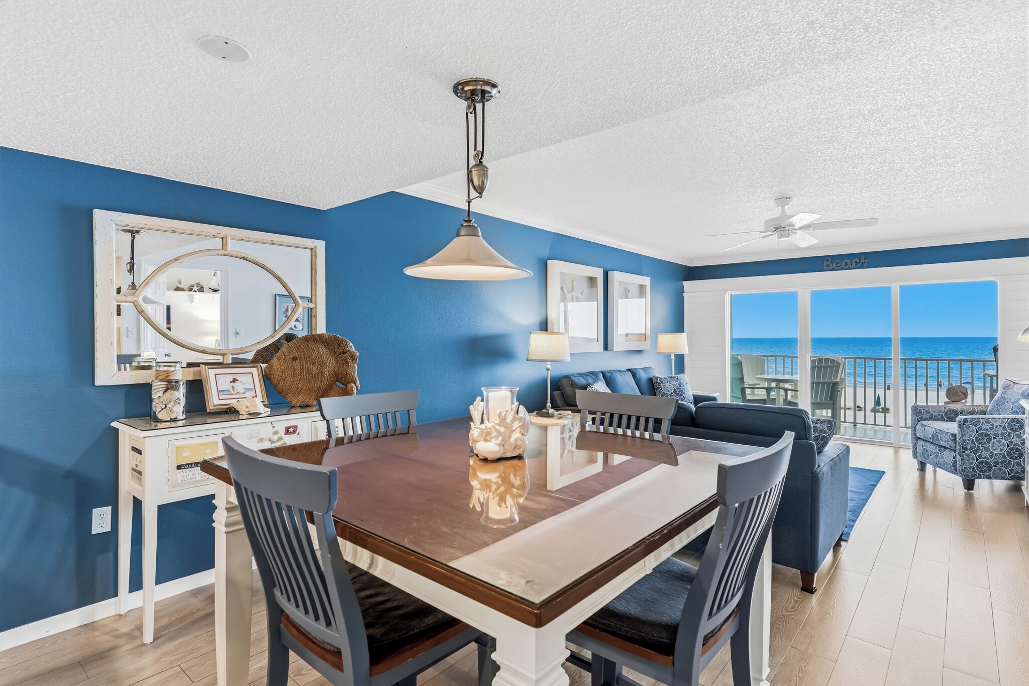 A dining area with a wooden table and chairs, a hanging light fixture, and a view of the ocean through large windows.