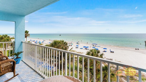 A balcony with wicker furniture, a potted plant, and a view of a beach with umbrellas and people.
