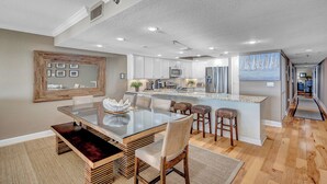 A modern kitchen with white cabinets, a central island with stools, and a dining area with a glass table and wooden chairs. - Huge 3rd Floor Gulf Front Condo w/ Balcony SD304 (Indian Shores)