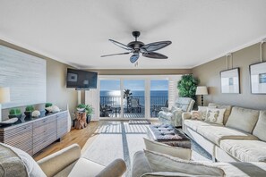 A spacious living room with a ceiling fan, a sofa set, a TV, and a balcony with a view of the ocean.
