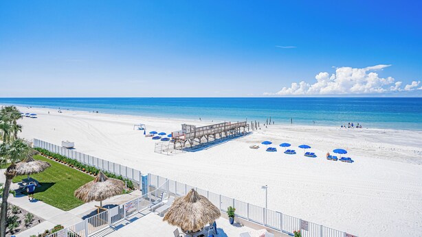 A beach with white sand, clear blue water, and a lifeguard tower. There are blue umbrellas and lounge chairs along the shore.