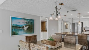 A modern dining area with a glass-top table, wicker chairs, and a painting of a lake scene. - Spacious Gulf Front Condo w/ Big Balcony - SD310 (Indian Shores)