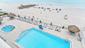A pool area with lounge chairs and a lifeguard stand, overlooking a beach with umbrellas and a pier.