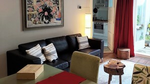Una sala de estar con un sofá de cuero negro, una mesa de centro de madera y un taburete pequeño de madera. Hay una obra de arte enmarcada en la pared y se ve una cocina a través de una puerta abierta.