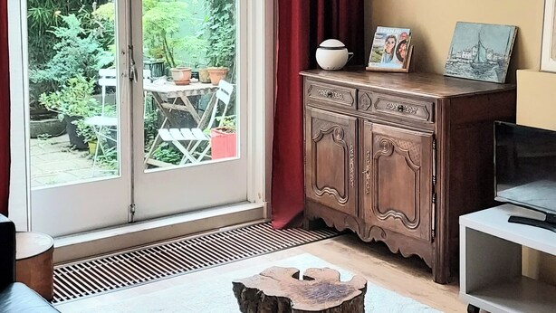 Un salón con puerta corrediza de vidrio a un jardín, un mueble de madera con marcos de fotos, un taburete pequeño y un televisor sobre un soporte.