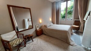 Un dormitorio con una cama grande, una silla de madera, una mesita de noche, un espejo y una ventana con cortinas.