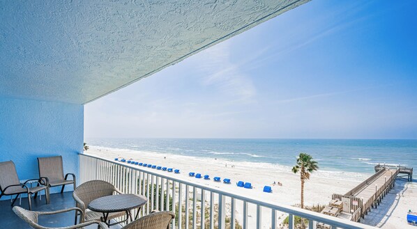 Ein Balkon mit Korbmöbeln, der auf einen Strand mit blauen Stühlen und einer Promenade blickt.