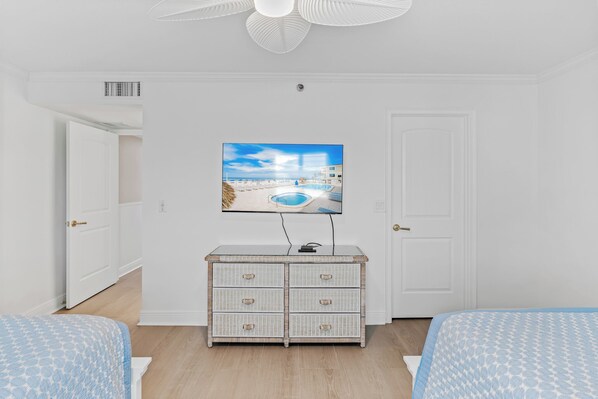 A bedroom with a TV mounted on the wall, a white dresser, and two beds with blue patterned bedding.