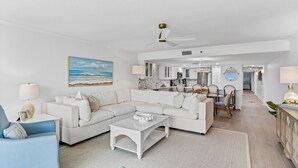 A spacious living room with a white sectional sofa, a blue armchair, and a coffee table. A ceiling fan and a painting of the ocean are visible.