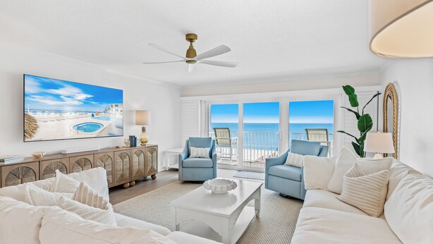 A spacious living room with a large window overlooking the ocean, a ceiling fan, and a view of the beach.