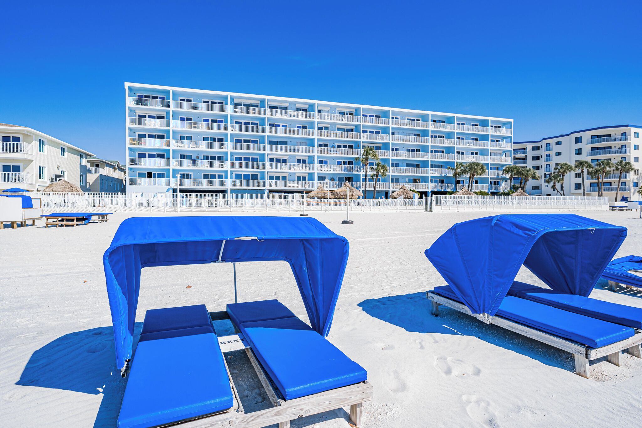 Un hotel frente a la playa con varios balcones, tumbonas con toldos azules y un cielo azul despejado.