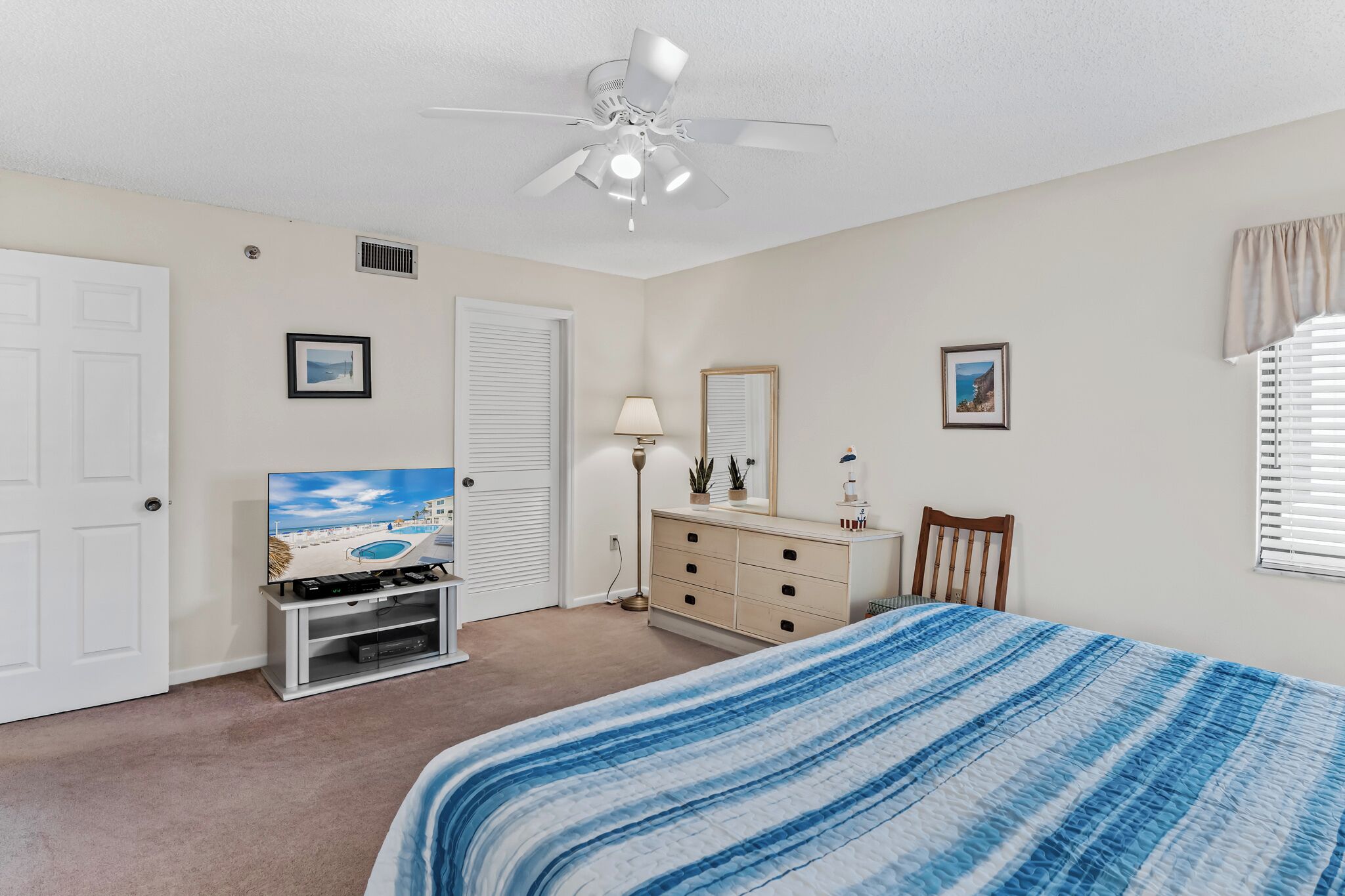 A bedroom with a bed, dresser, TV, ceiling fan, and a window with blinds.