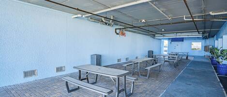 A covered outdoor area with picnic tables and benches, a blue wall, and a ceiling with exposed pipes.