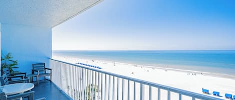 A balcony with a view of a beach, a table, and chairs.