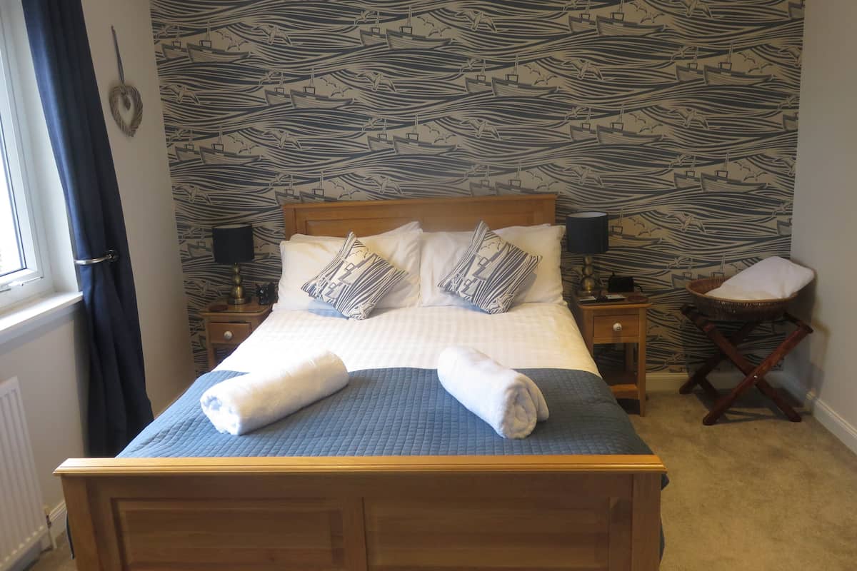 Un lit bien fait, recouvert d’une couette bleue, avec deux oreillers à motifs marins et une tête de lit en bois. On y trouve une table de chevet avec une lampe, ainsi qu’une petite table sur laquelle est posé un panier.