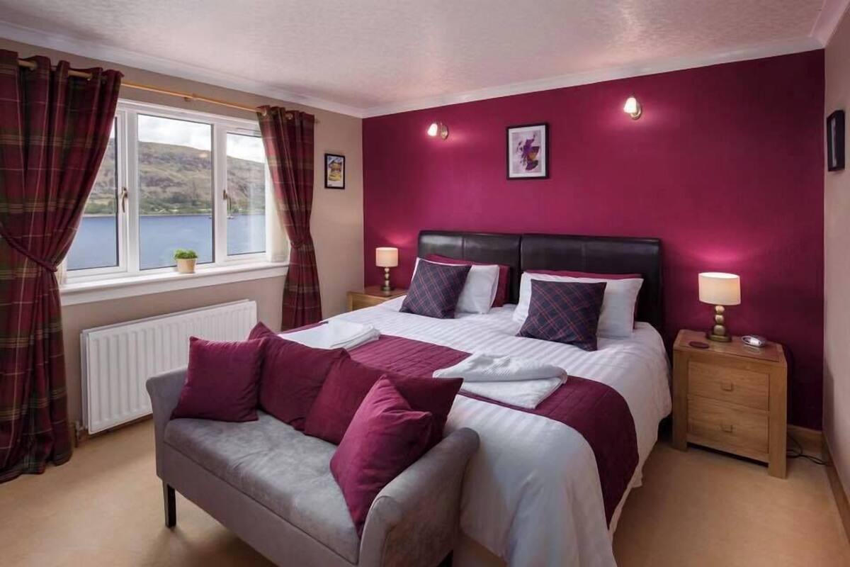Une chambre à coucher comprenant un lit, une table de chevet, une lampe, une fenêtre avec des rideaux et une vue sur l’extérieur.