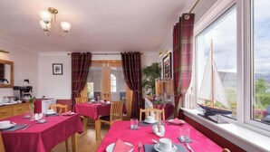 A dining area with tables set for four, each with a red tablecloth, white plates, and cutlery. There is a window with a view of greenery and a sailboat model. - Blythedale House (Fort William)