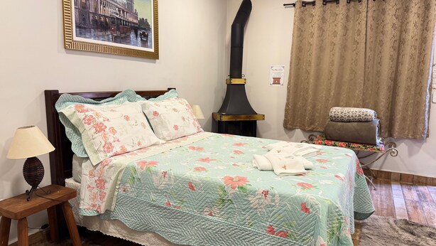 A neatly made bed with floral bedding, a nightstand with a lamp, a framed picture on the wall, and a large chimney.