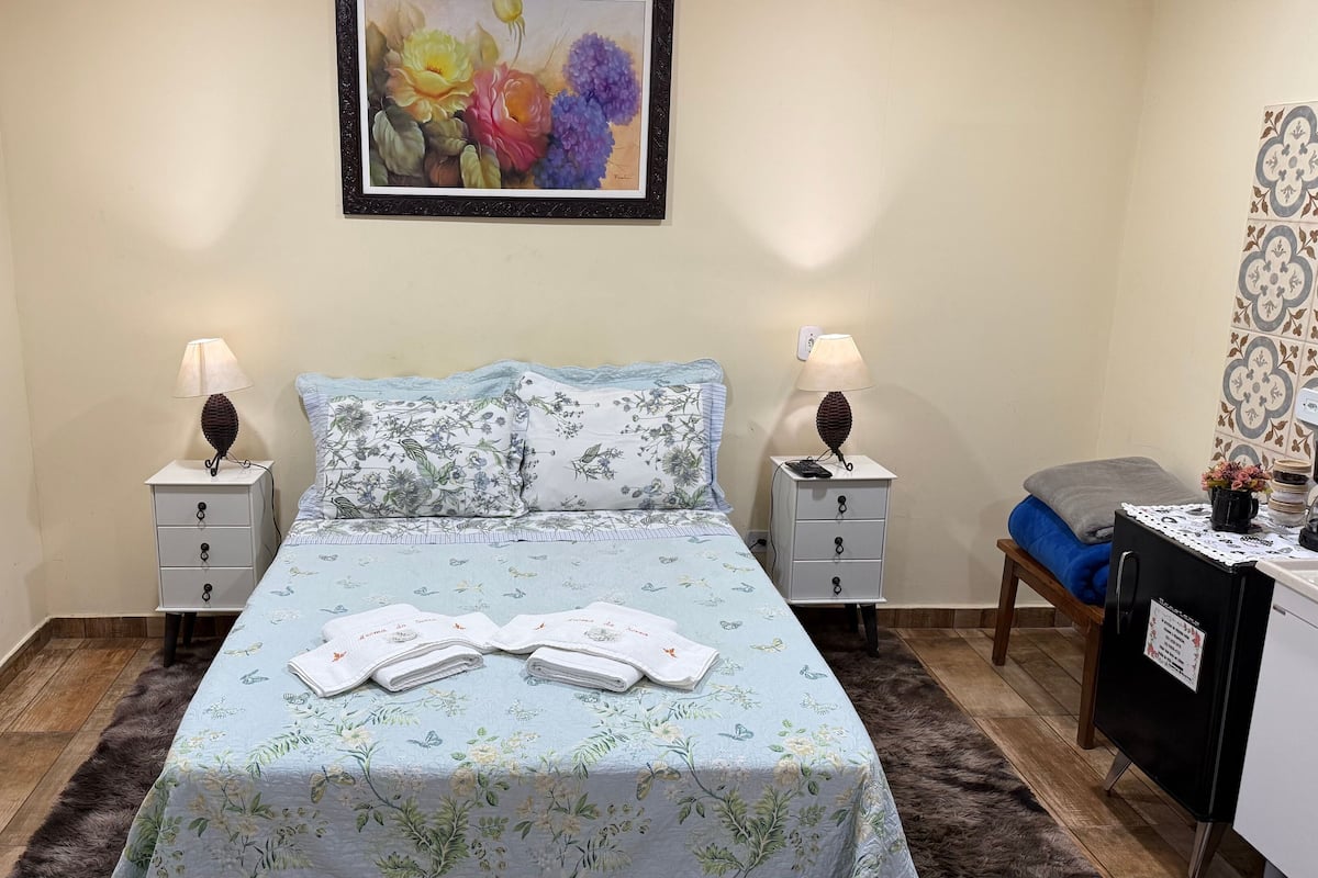 A bedroom with a bed, two bedside tables with lamps, a desk with a chair, a painting on the wall, and a rug on the floor.